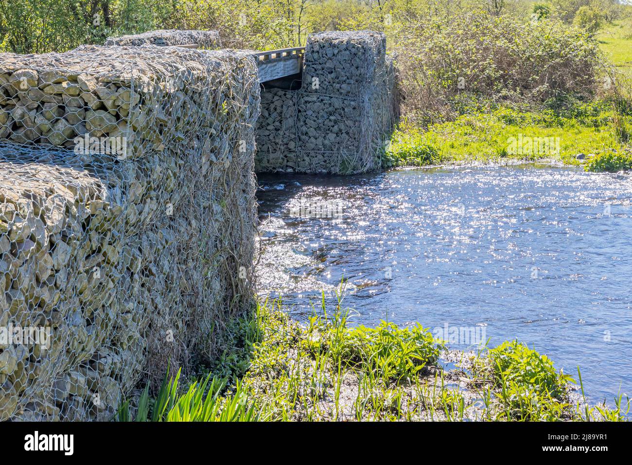 Wire gabion rock wall, part of a pedestrian bridge over a river with wild vegetation in the background. New ecological architecture in the manufacture Stock Photo