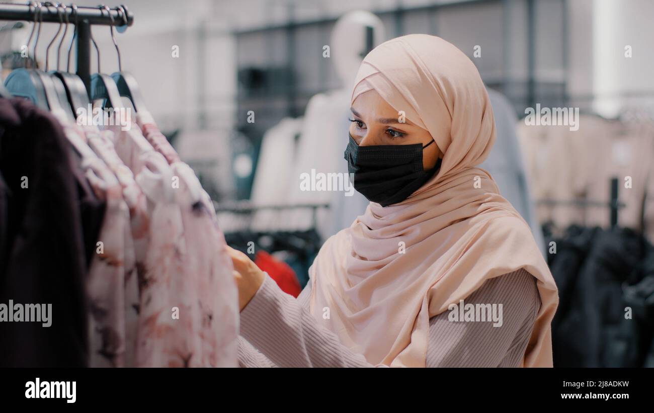 Young arab woman customer buyer in hijab wearing protective medical mask in clothing store carefully chooses clothes in search suitable outfit looking Stock Photo