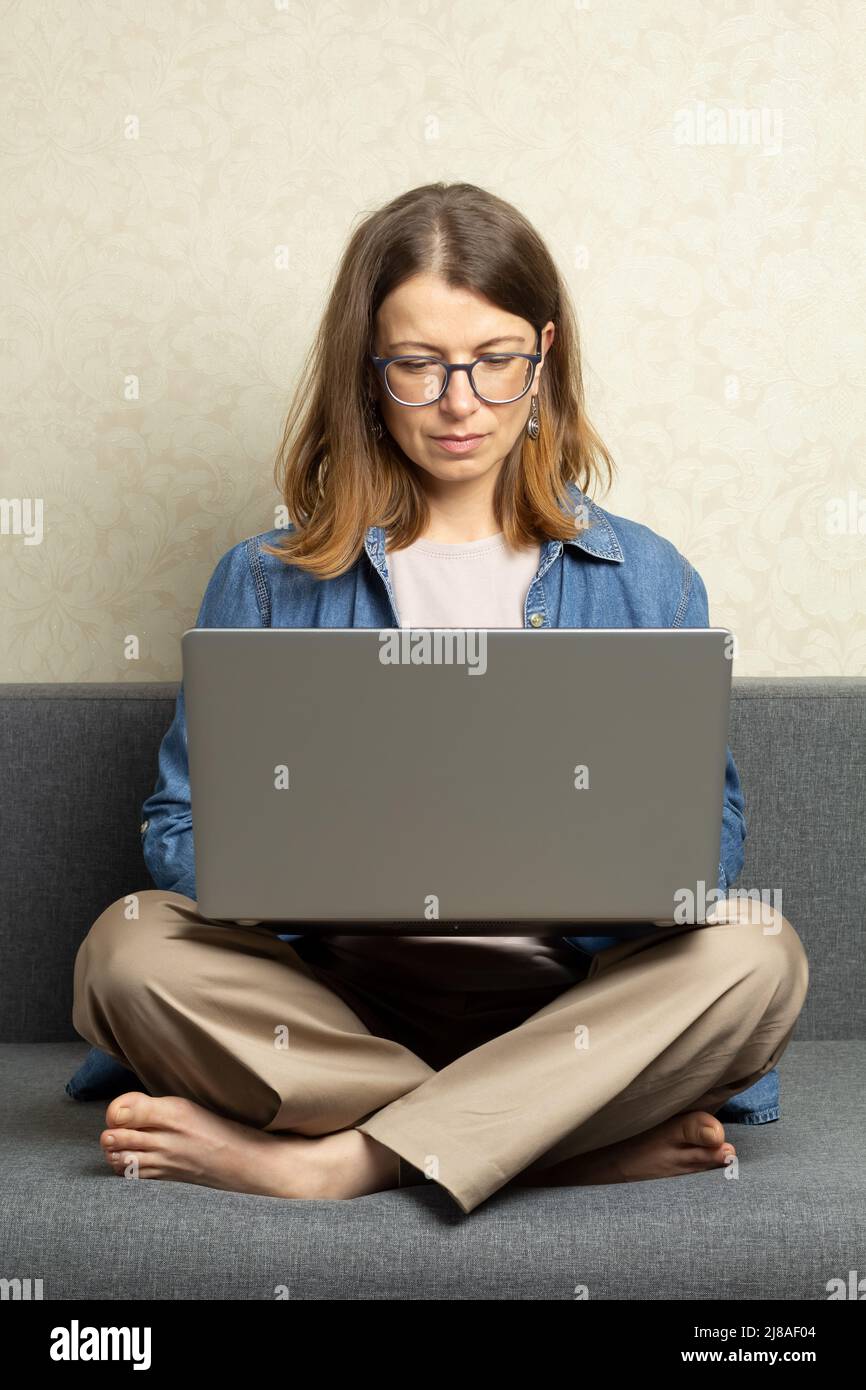 A young woman works on a laptop while sitting on the couch at home. Stock Photo
