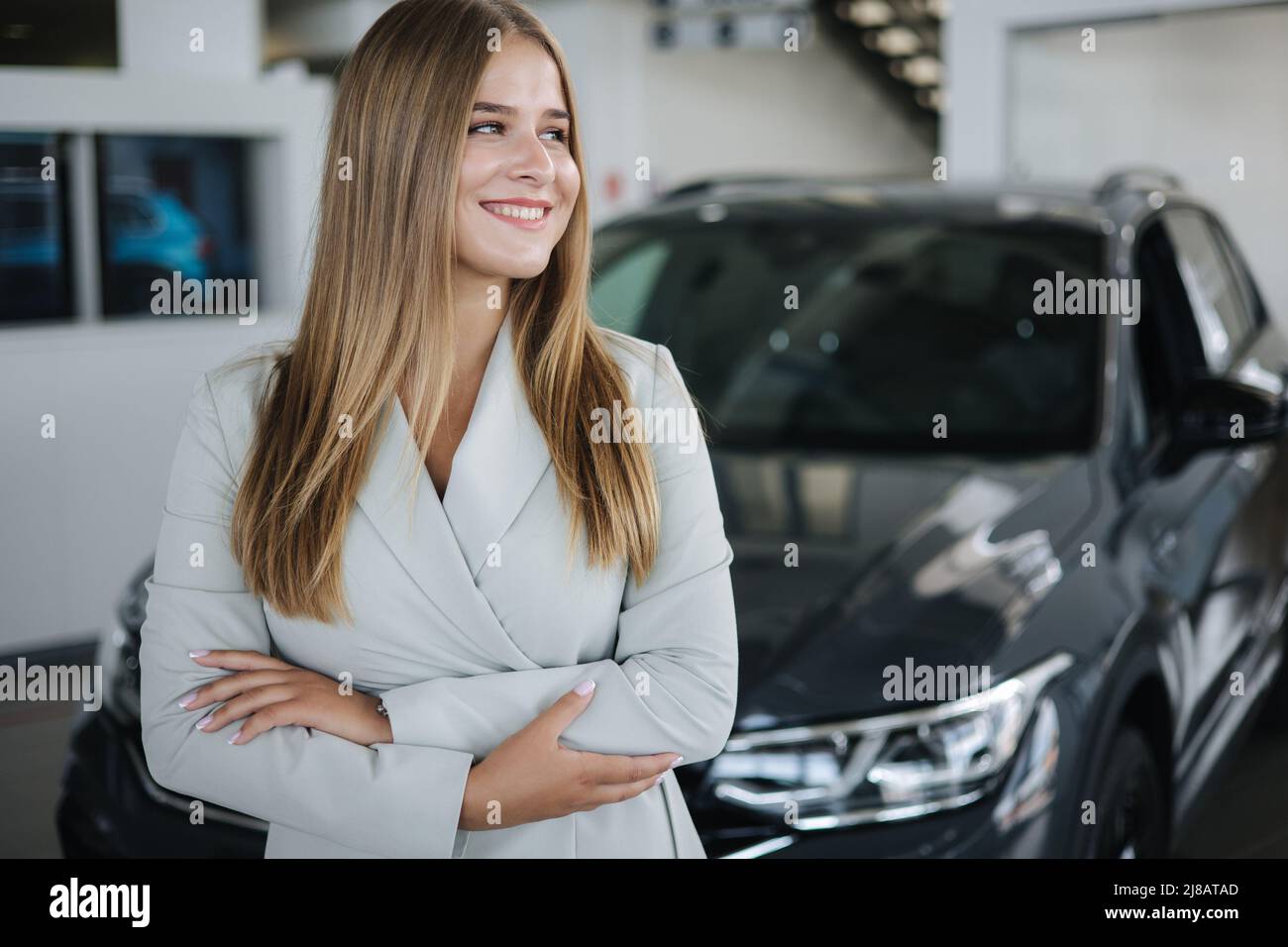 Portrait of salesperson in car showroom. Attractive young woman stend in front of car. Beautiful woman in suit in front of luxury car. Stock Photo