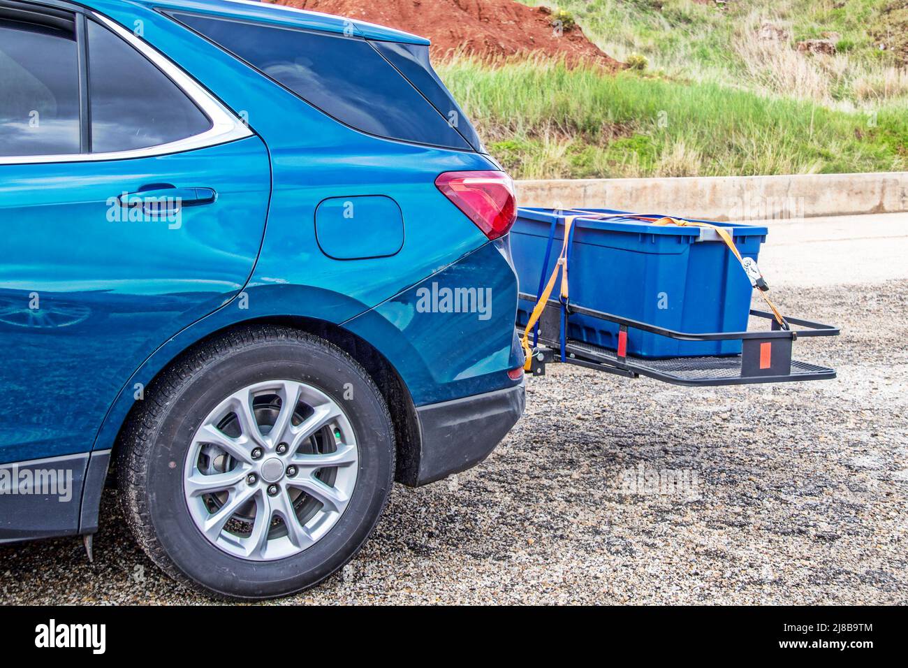 Blue hatchback car parked on gravel with hitch cargo carrier holding tied down rubber storage box on back - Closeup and cropped Stock Photo