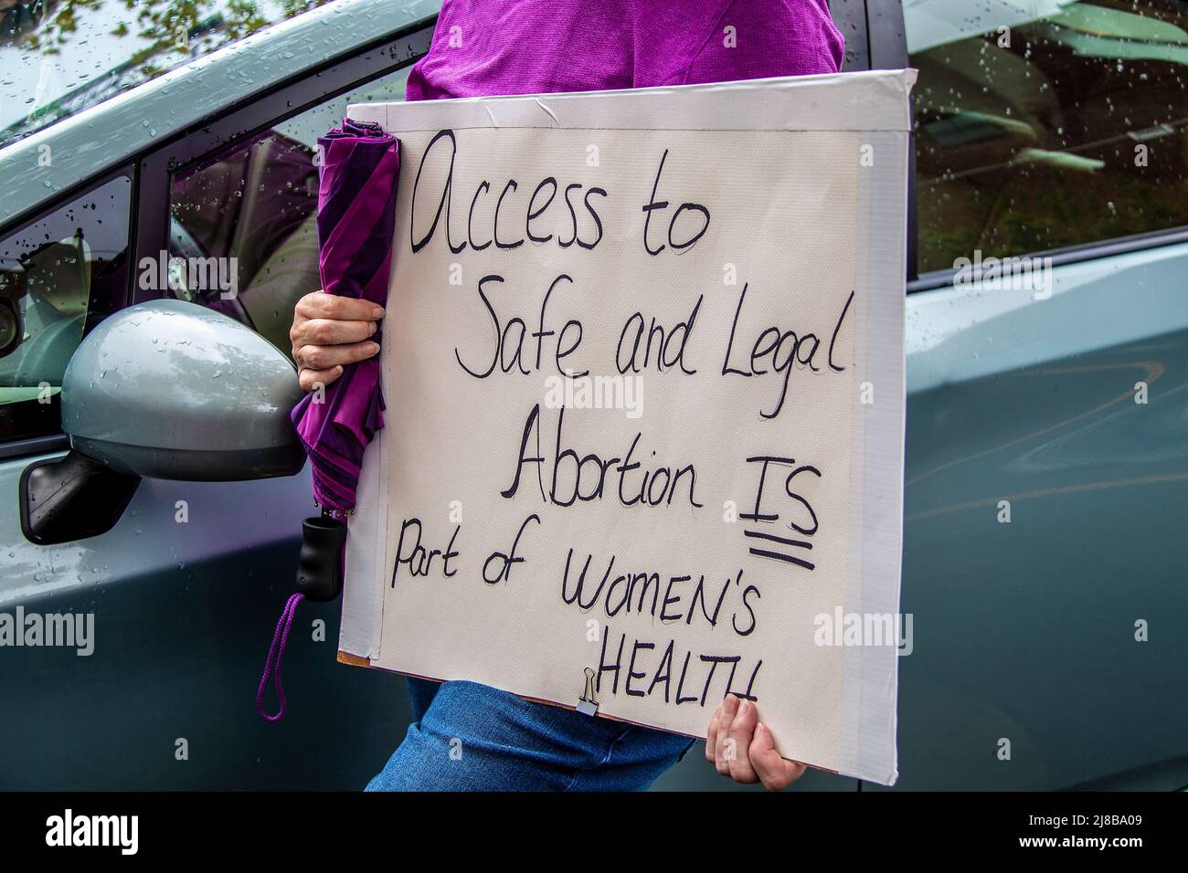 Home made sign reading Access to Safe and Legal Abortion Is part of womens health held by cropped woman in purple shirt holding umbrella standing by c Stock Photo