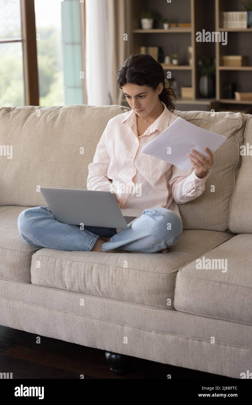 Serious woman sitting on sofa holds documents working on computer Stock Photo