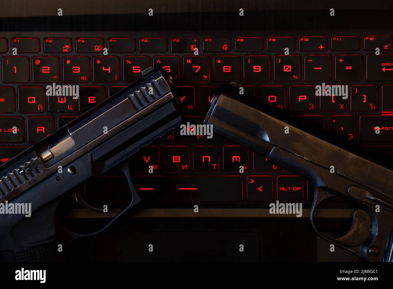 Two guns lie on a black laptop keyboard with red backlight, security threat, cyber attack, system hack Stock Photo