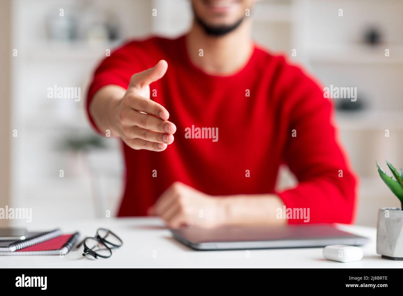 Happy friendly young arabic guy gives hand welcoming at camera at workplace in home office interior, blurred Stock Photo