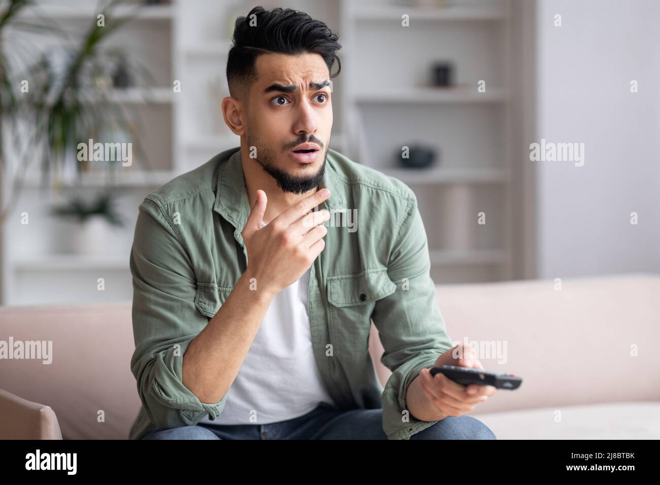 Shocked frightened young arab man with beard with remote control watches news on TV sits on sofa Stock Photo