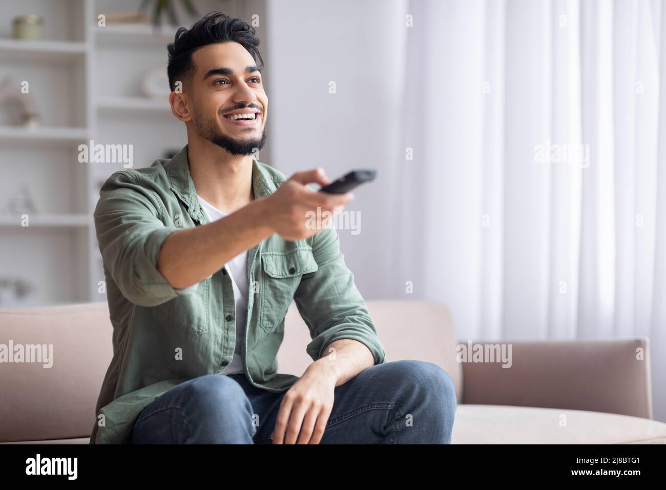 Smiling islamic guy with beard sits on sofa with beard with remote controls watches match on TV, sits on sofa Stock Photo
