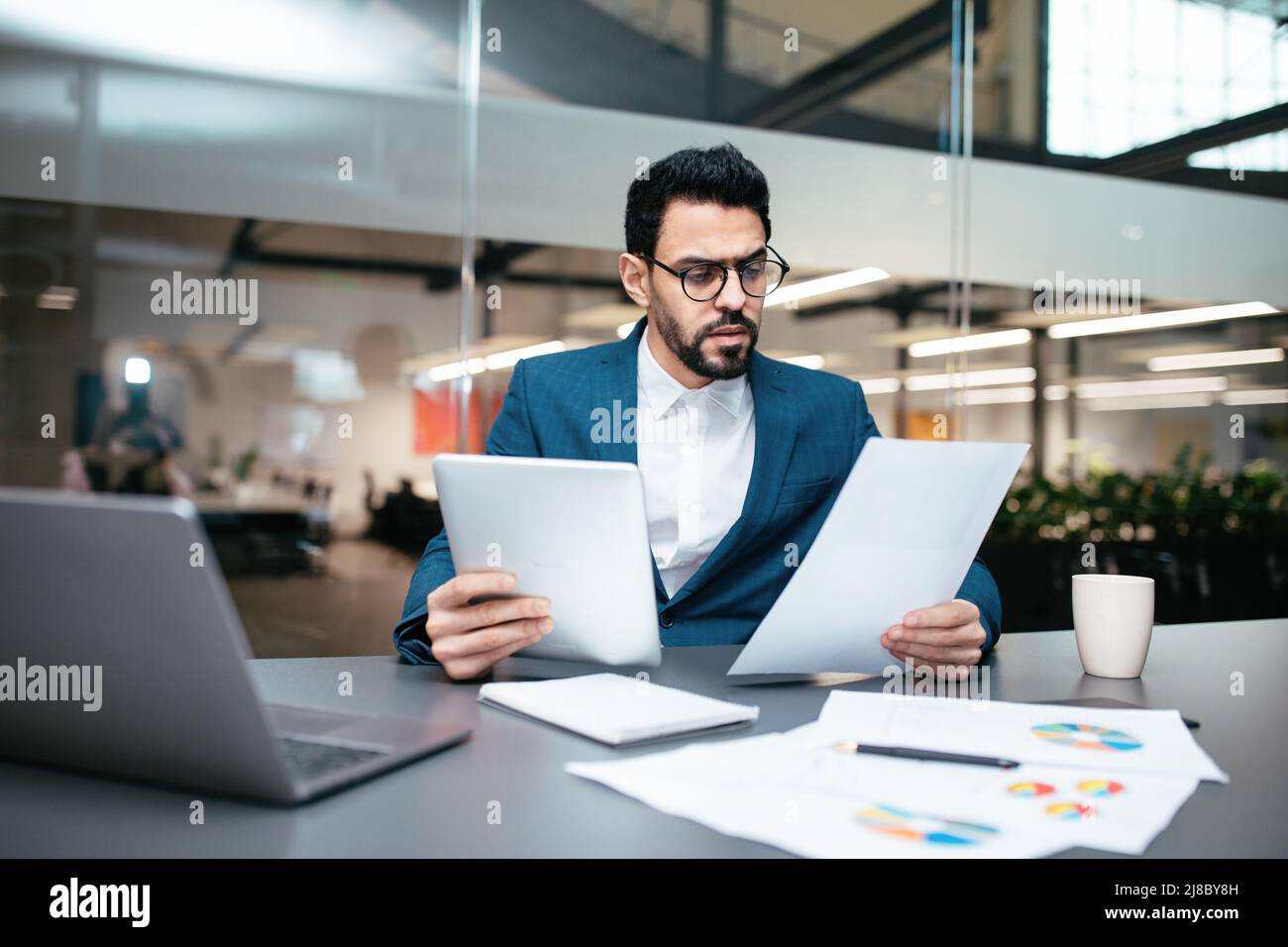 Serious busy handsome young muslim guy with beard in glasses, suit sits at table with computer and tablet Stock Photo