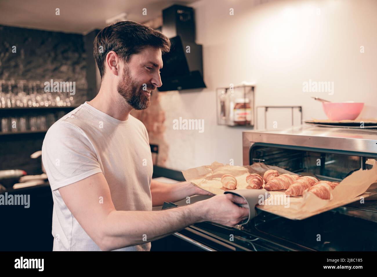 Caucasian young handsome baker taking out croissants out of oven and smiling. Bakery concept. Stock Photo
