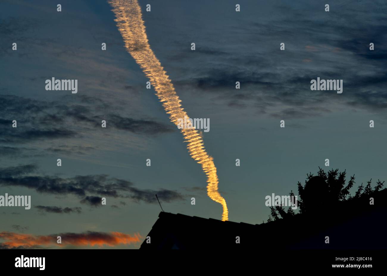 single appealing and luminous condensation trail in bright light orange color above silhouettes of roofs in the dark blue evening sky (red cloud) Stock Photo