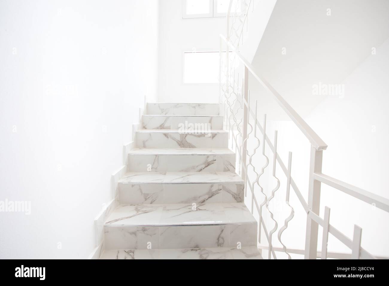 A bright, modern staircase. Stairs go up where you can see windows and light. White metal handrail on the side. Stock Photo