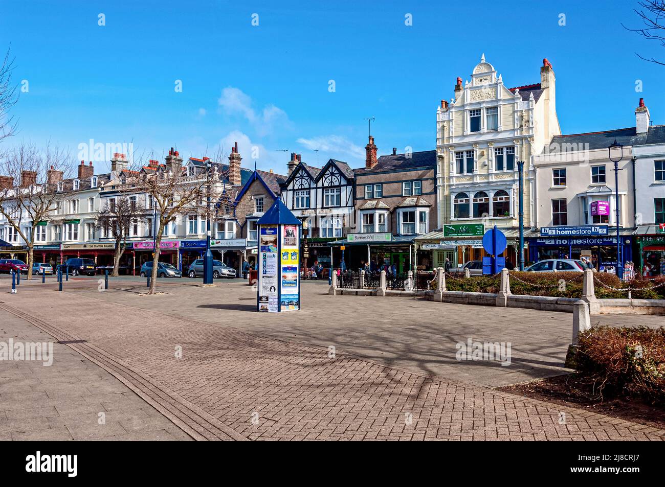 A peaceful paved square with a herring bone patterned strip and shrubbery beds faces a parade filled with interesting shops and other businesses Stock Photo