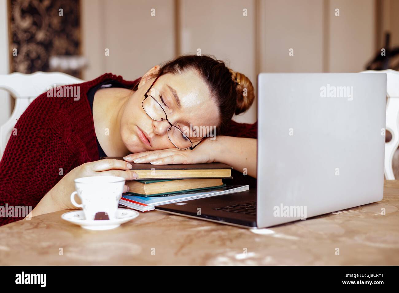Tired young woman in glasses sleeping on books near laptop at desk in home room. Tiredness from monotonous study and rest at workplace from computer Stock Photo