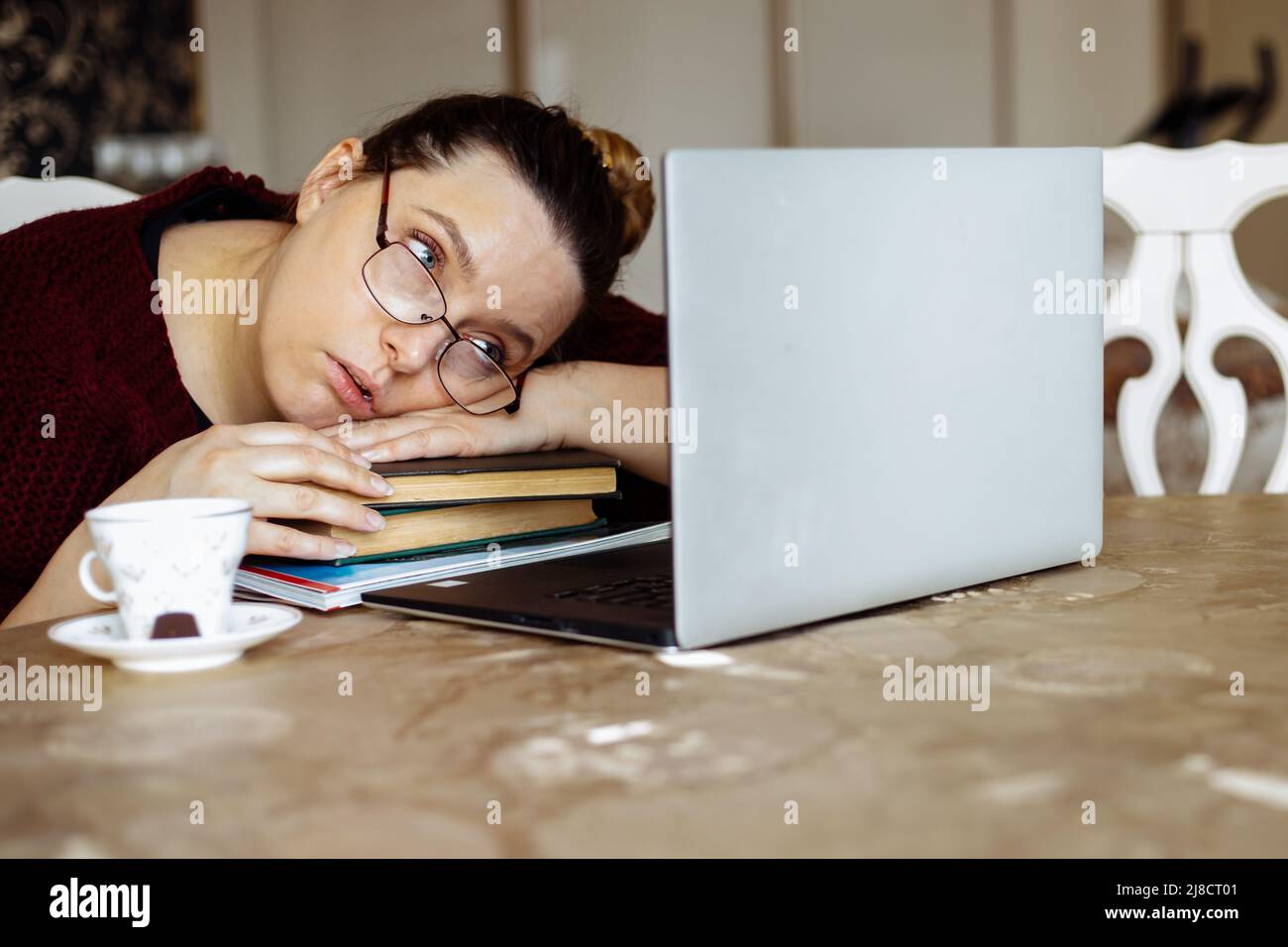Tired young woman in glasses lying down on books and looking at laptop screen at desk in home room. Tiredness from monotonous study and computer Stock Photo