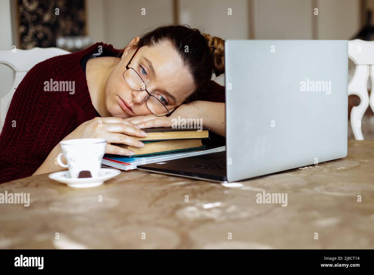 Tired young woman in glasses lying down on books and looking away near laptop at desk in home room. Tiredness from monotonous study and computer Stock Photo