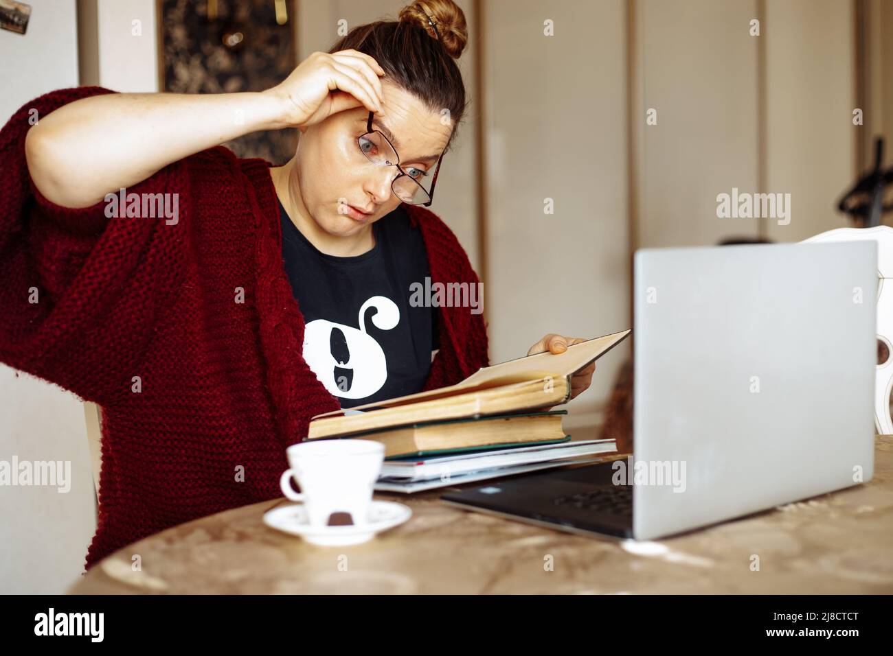 Tired young woman taking off glasses sitting at desk in home room near laptop and reading book. Tiredness from monotonous study and computer overwork Stock Photo