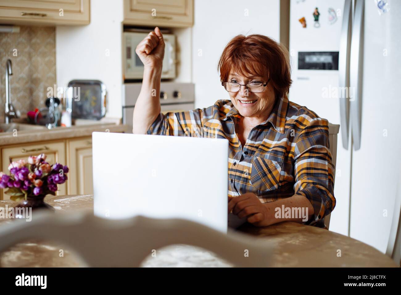 Happy mature lady looking at laptop screen sitting at table in kitchen. Advanced pensioner practicing computer skills and learning modern technology Stock Photo