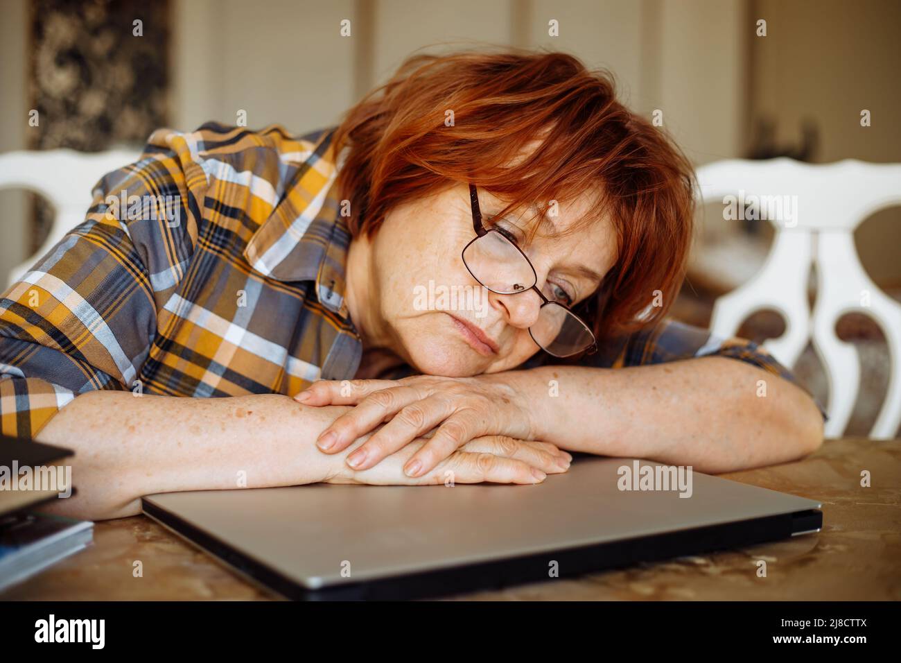Tired mature lady in glasses lying down on laptop at desk in home room. Advanced pensioner practicing computer skills and learning modern technology Stock Photo