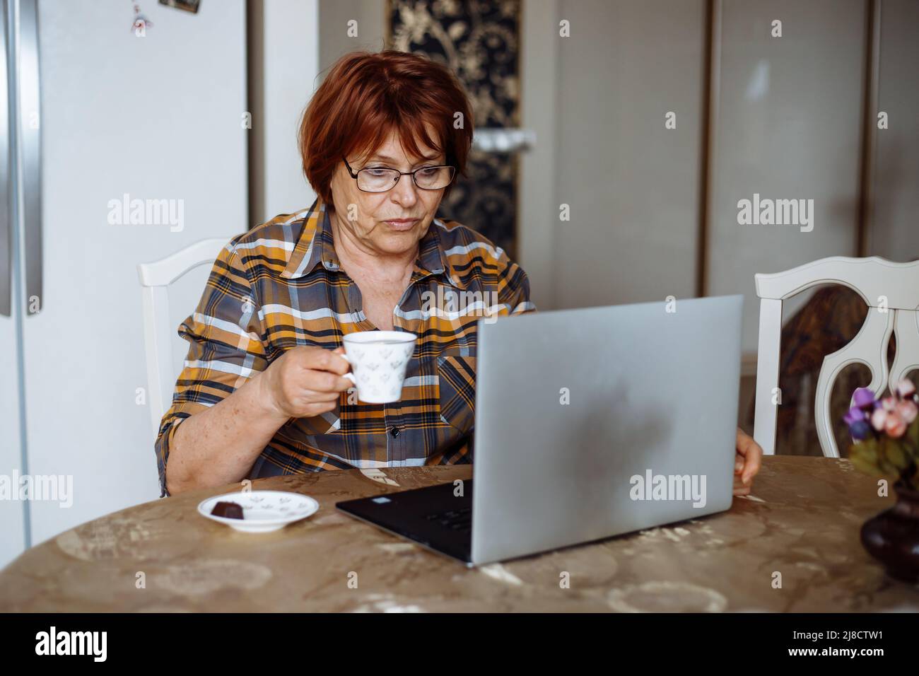 Mature lady in glasses looking at laptop screen at table in kitchen and holding mug of drink. Advanced pensioner practice and learn modern technology Stock Photo