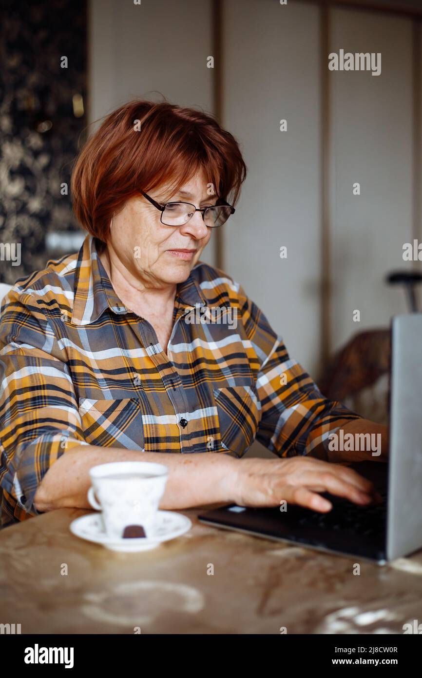 Mature lady in glasses typing on laptop at desk in room with mug of drink. Advanced pensioner practice computer skills and learn modern technology Stock Photo