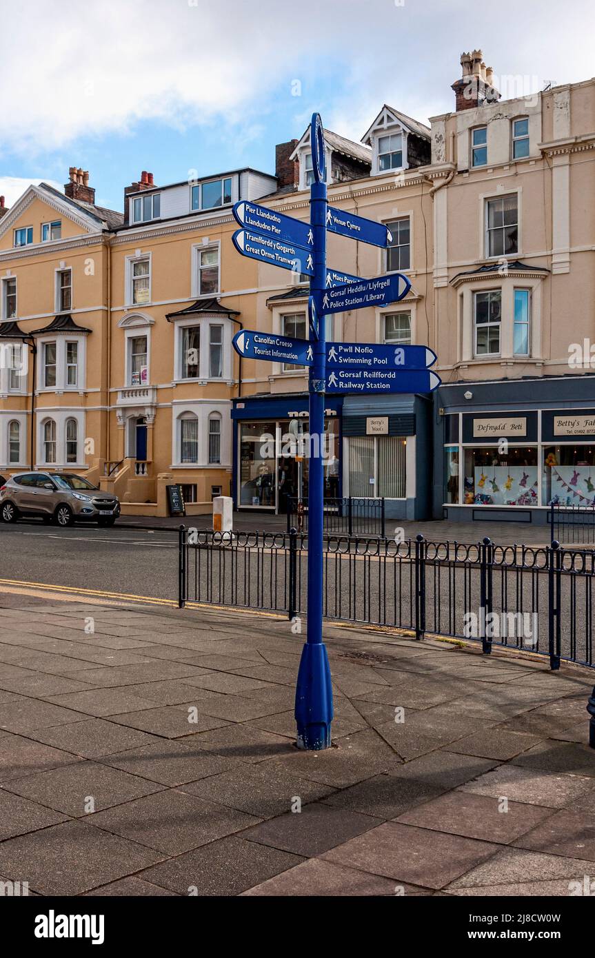 A bilingual signpost with directions printed in the Welsh and English languages stands behind metal  railings at the corner of a road in Llandudno Stock Photo