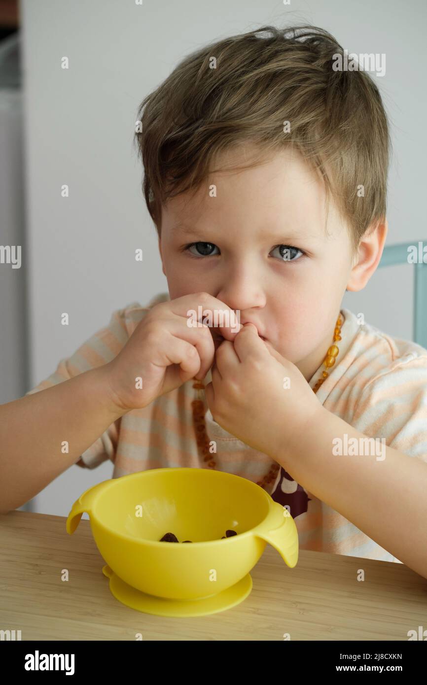 Toddler boy eating happily by the table food by himself in the room. Child having meal. Happy kid having lunch. Little kid with bowl Stock Photo