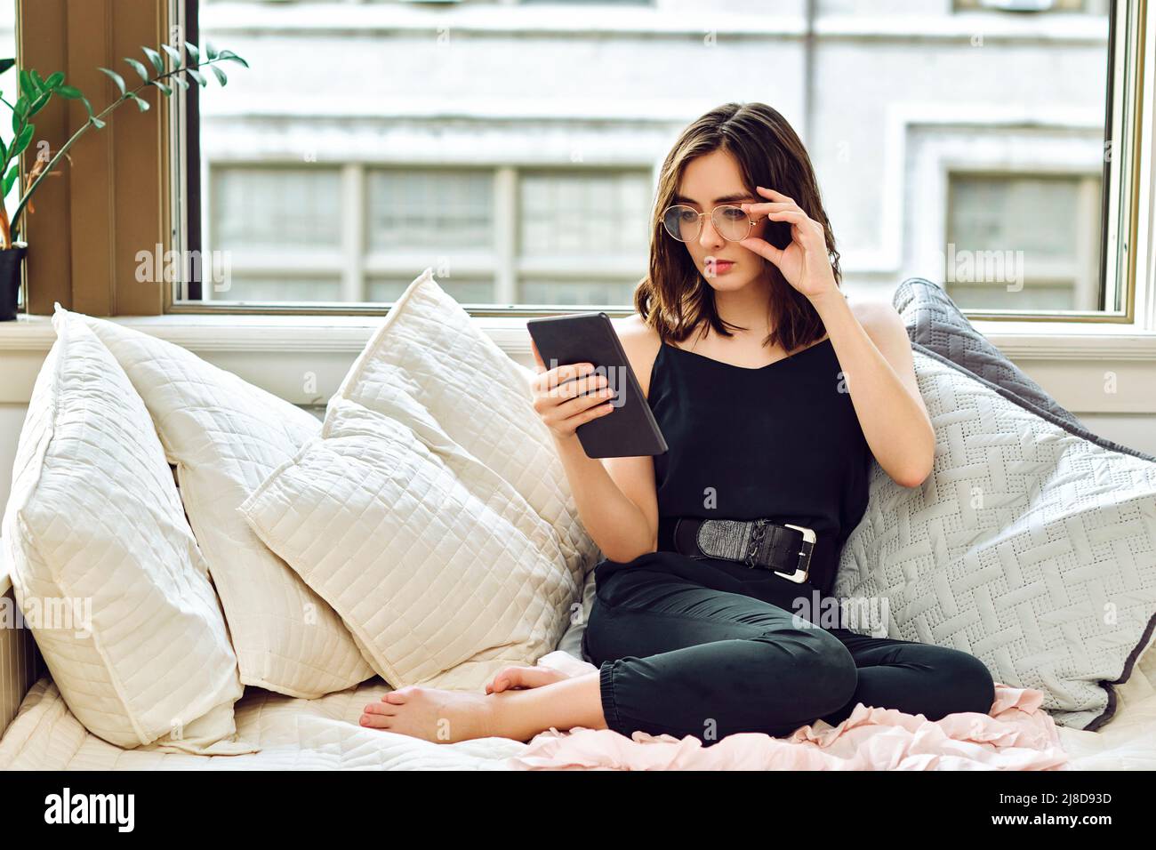 thoughtful beautiful young adult woman with eyeglasses reading tablet on sofa indoors Stock Photo