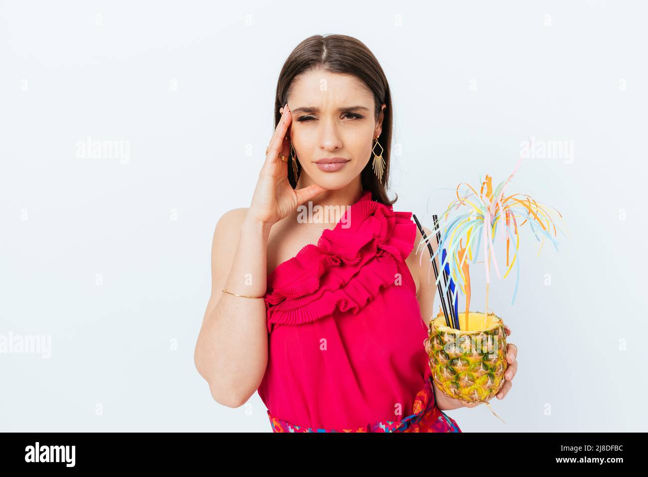 Beautiful fashionable young woman holding festive pineapple cocktail tiredly holding her head squinting her eyes Stock Photo
