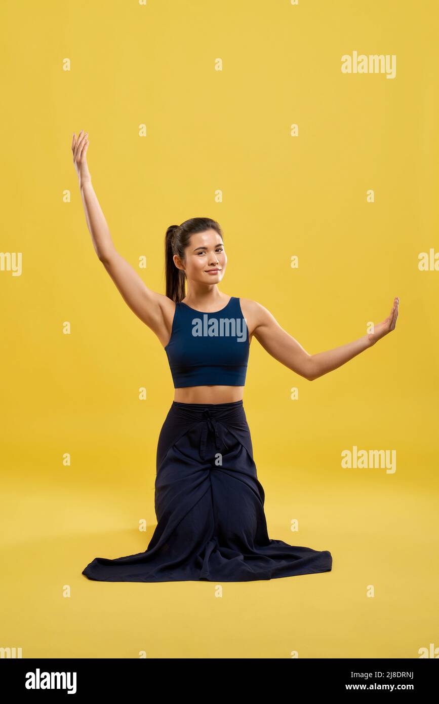 Front view of sporty female practicing yoga pose indoors. Brunette with pony tail standing on knees, raising hands, looking at camera. Isolated on yellow studio background. Stock Photo