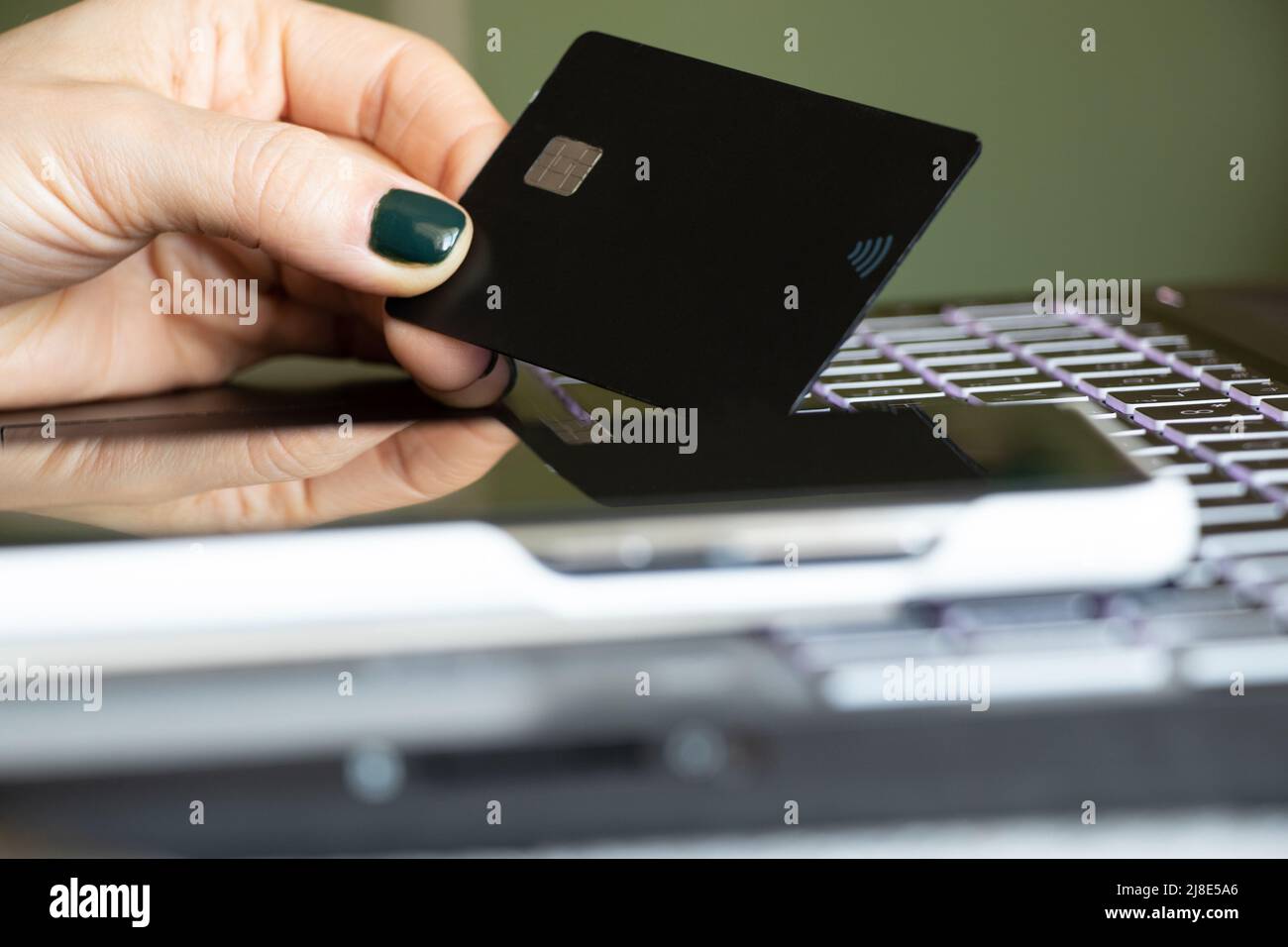 A girl holds a credit bank card in her hands next to a black laptop and a phone in the office by the window, online payment, payment by card Stock Photo