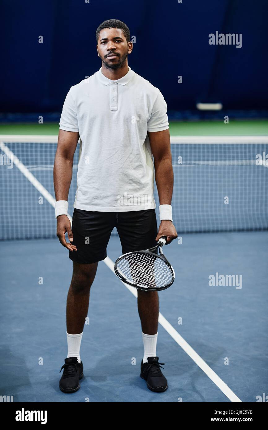 Full length portrait of African American tennis player looking at camera while posing confidently with racket at indoor court Stock Photo