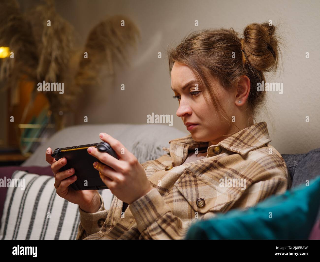 A gamer girl is looking intently at the screen of a portable game console, sitting on a sofa in a room. Video games, mobile games, virtual reality, es Stock Photo