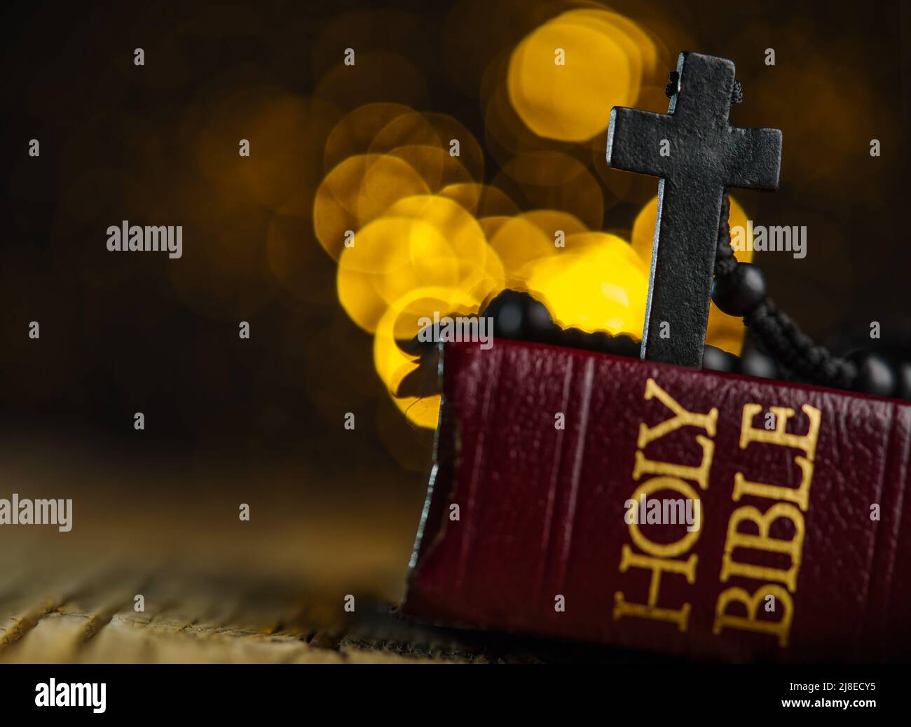 On a simple wooden table lies a red book - the Holy Bible and on it a black Catholic cross on a rosary against a yellow background with twinkling ligh Stock Photo