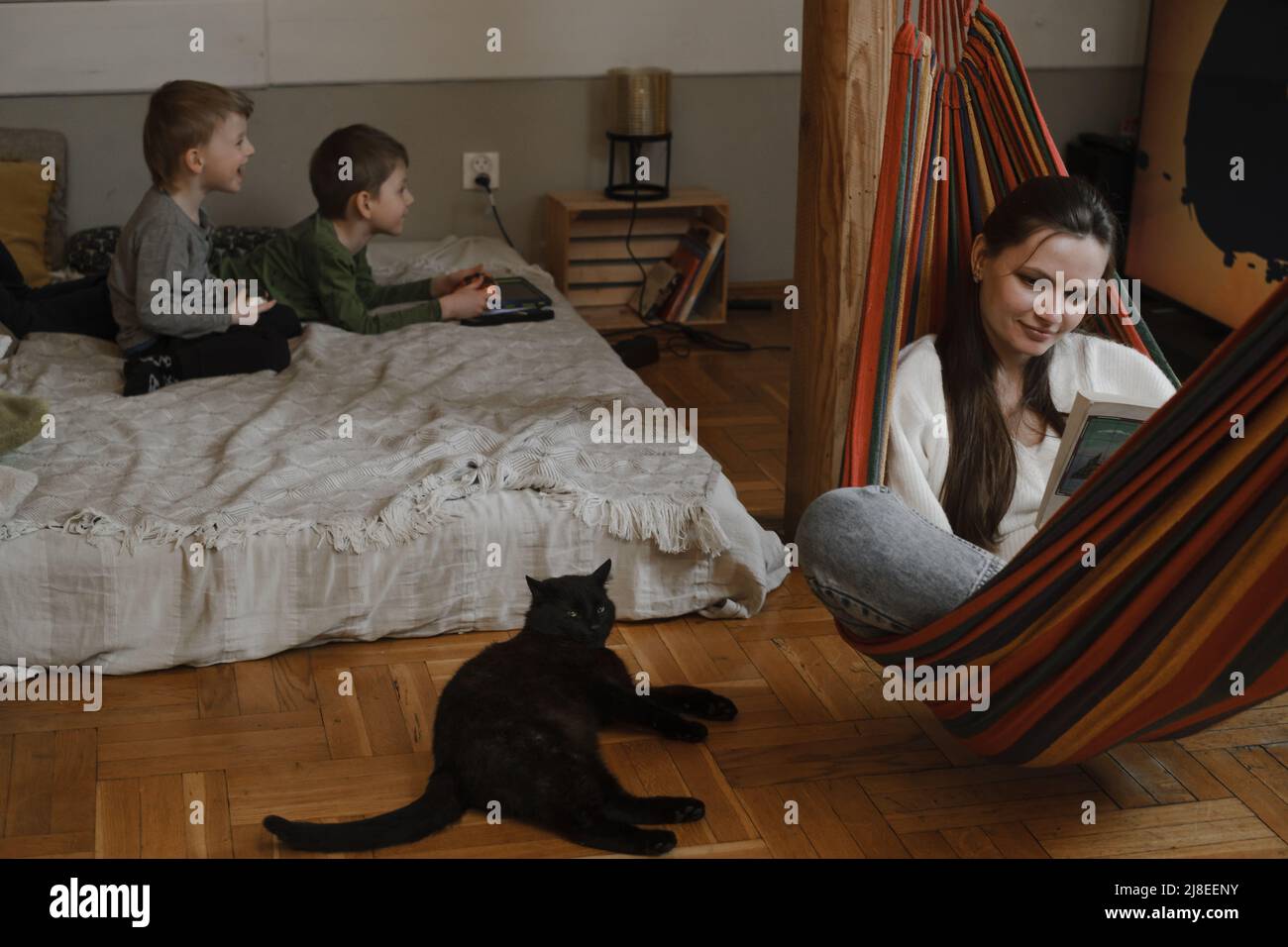 Kids are playing play station while mother is reading book in hammock. Family with black cat as a pet. Brothers playing video games on the bed. Family Stock Photo