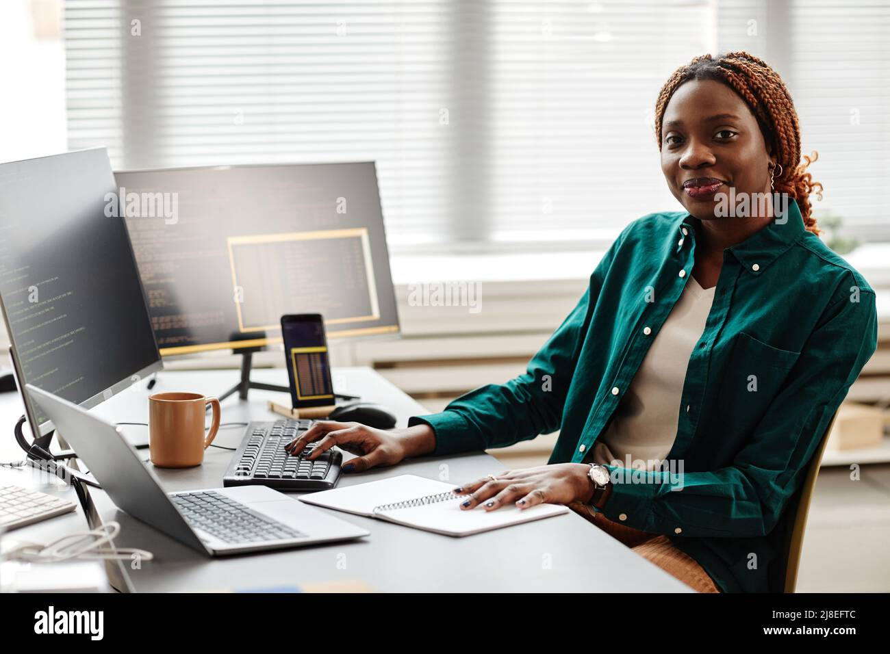Warm toned portrait of black young woman as IT developer or QA engineer smiling at camera while sitting at workplace with computers, copy space Stock Photo