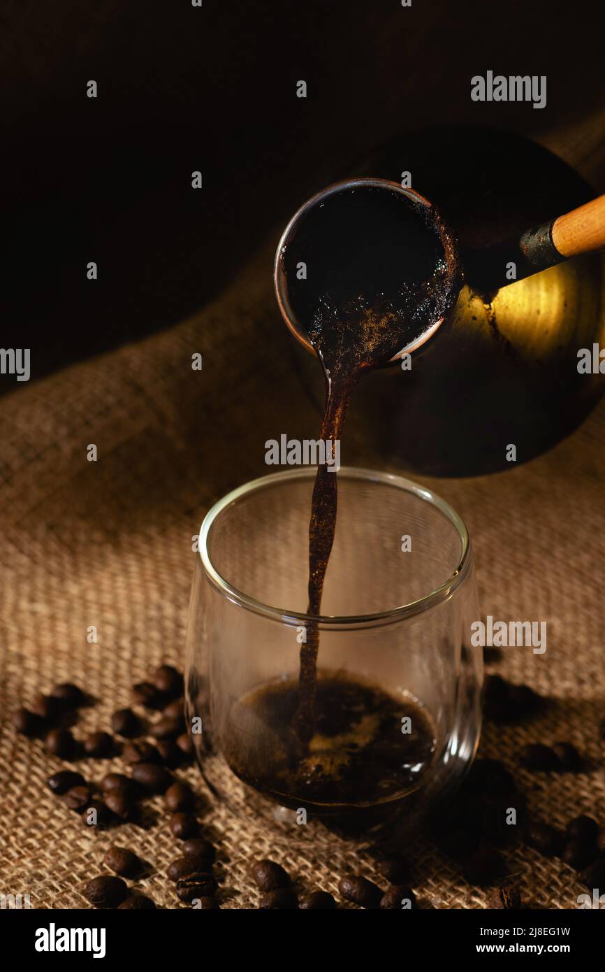 Freshly brewed espresso coffee is poured from a cezve into a transparent glass cup against a dark background of burlap with coffee beans Stock Photo