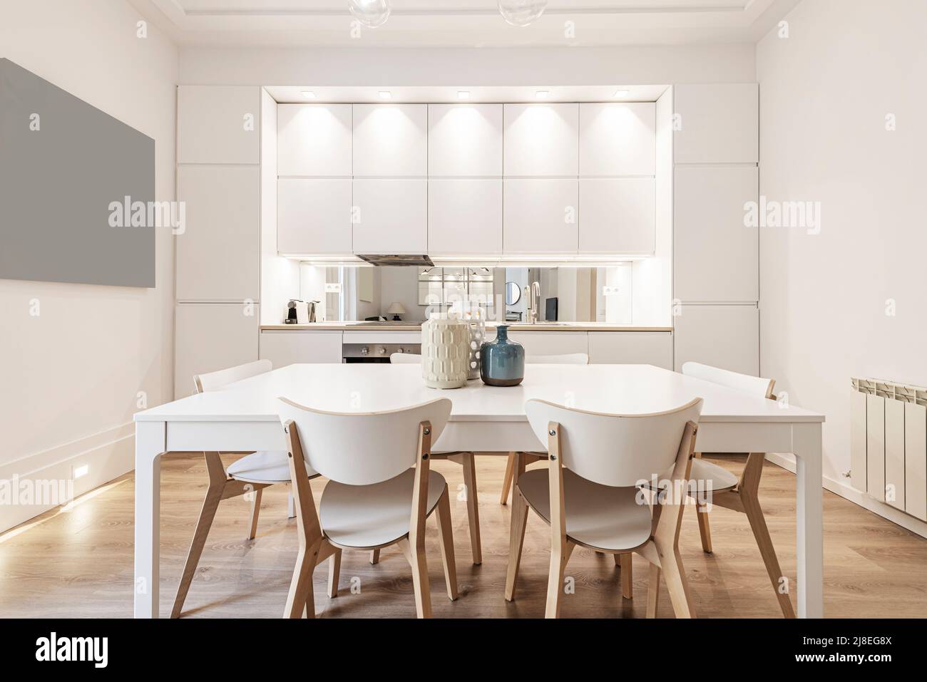 White wooden dining table with matching wooden chairs in front of a kitchen with plain white