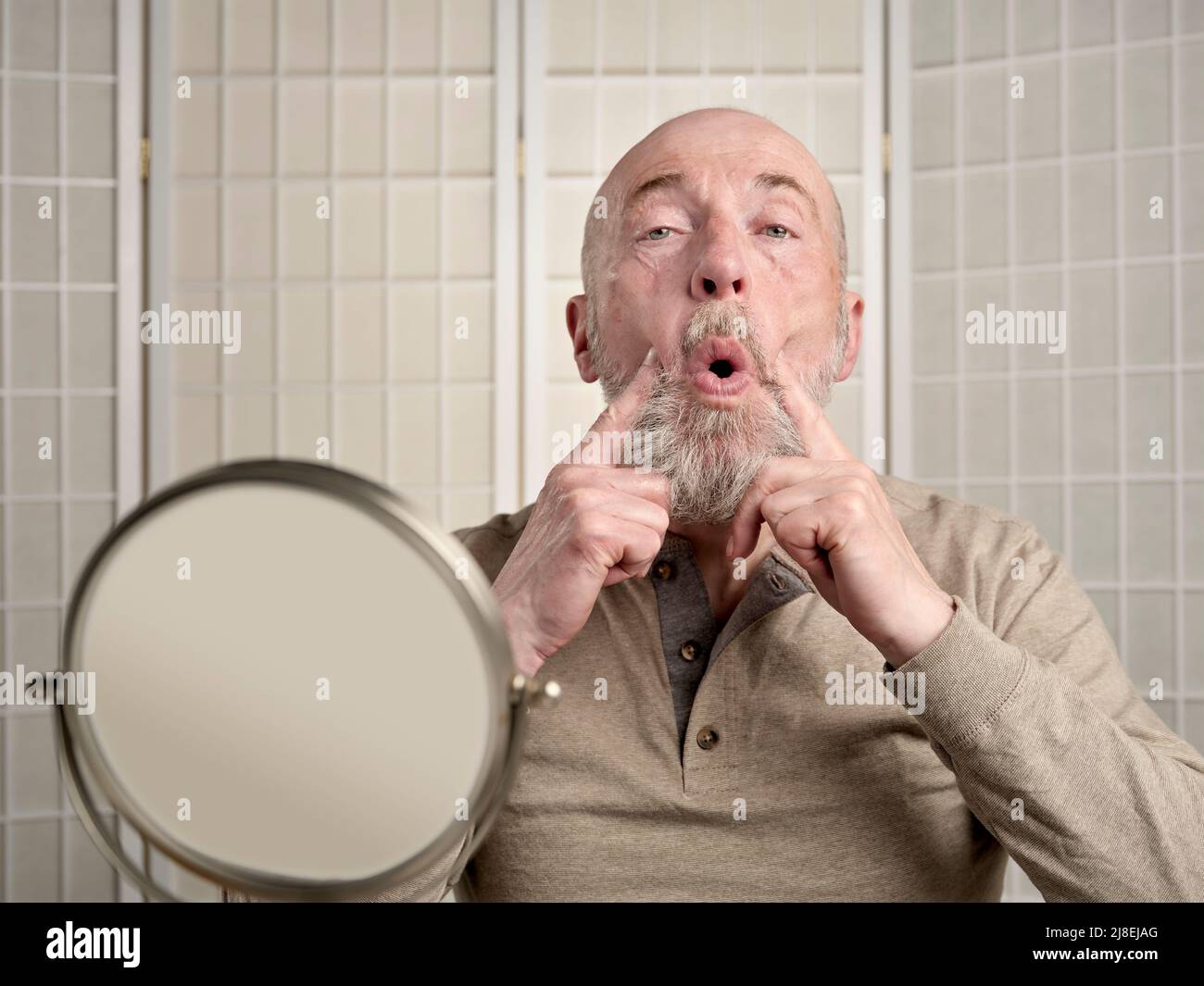 Head and shoulders portrait of a bald, bearded senior man performing face toning exercise (eye toner), self care concept Stock Photo