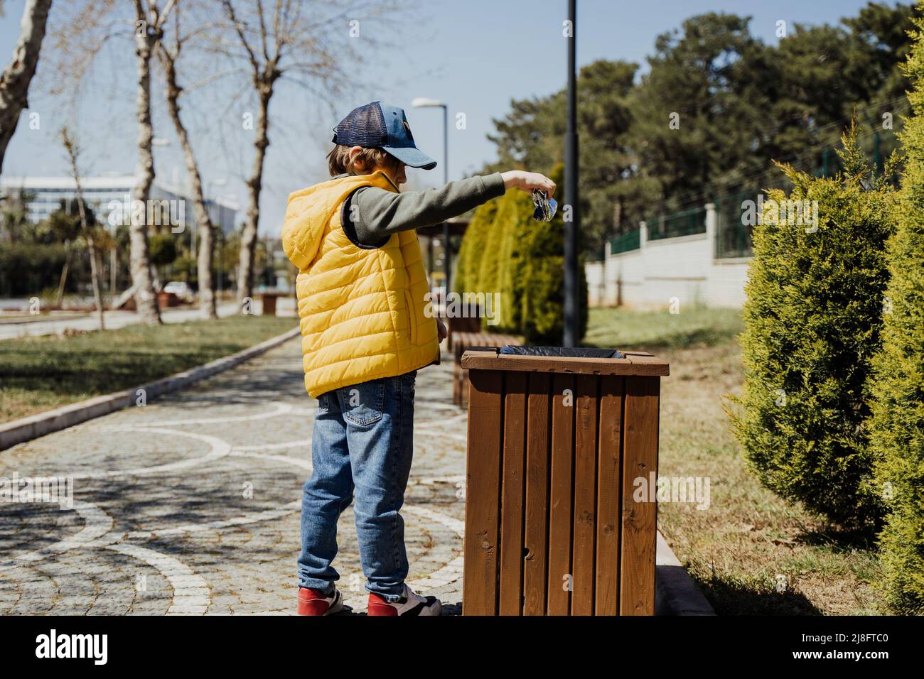 Schoolboy kid throwing the trash into dumpster. Boy using recycling bin to throw away the litter. Caucasian child recycles the junk into the trash-can Stock Photo