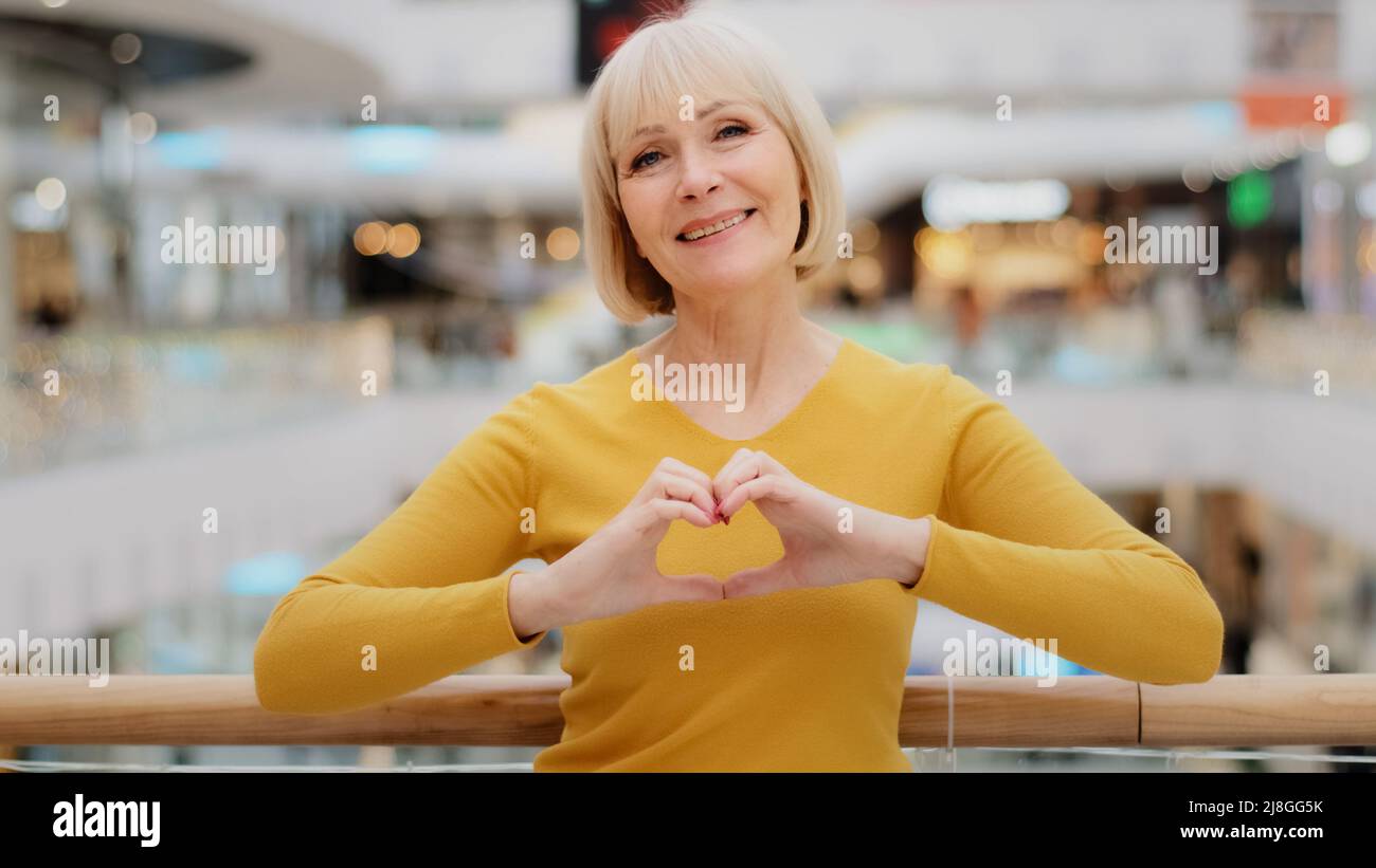 Mature happy caucasian woman standing in public place posing sincerely smiling looking at camera making heart shape with fingers sign generosity and Stock Photo