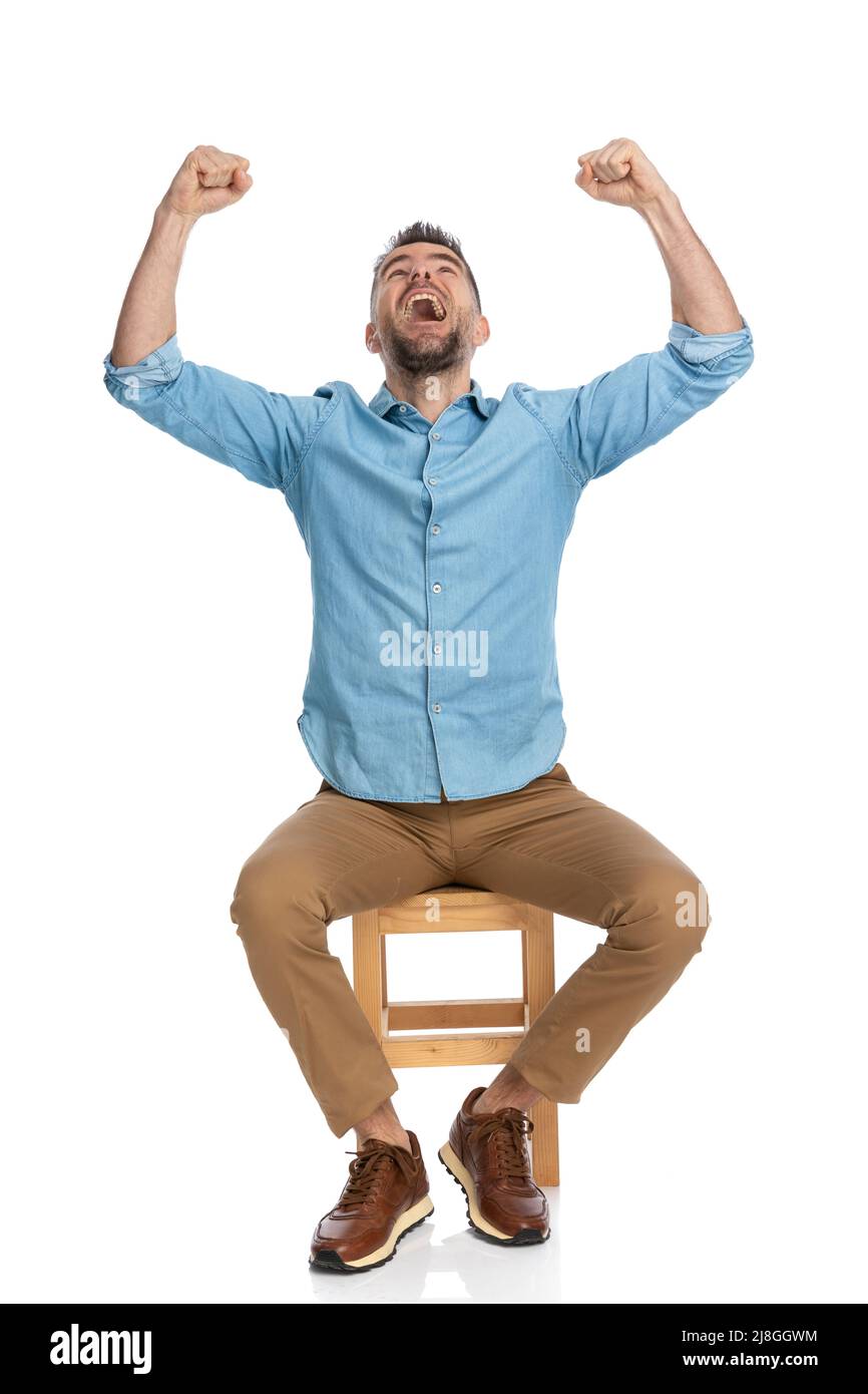 excited man in his forties with arms above head looking up, yelling and celebrating victory while sitting on wooden chair in front of white background Stock Photo