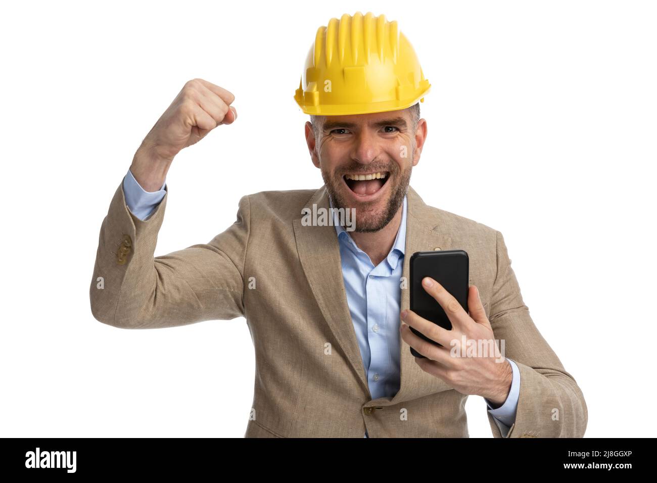 handsome bearded engineer guy in suit with helmet holding telephone, laughing with arm in the air and cheering in front of white background Stock Photo