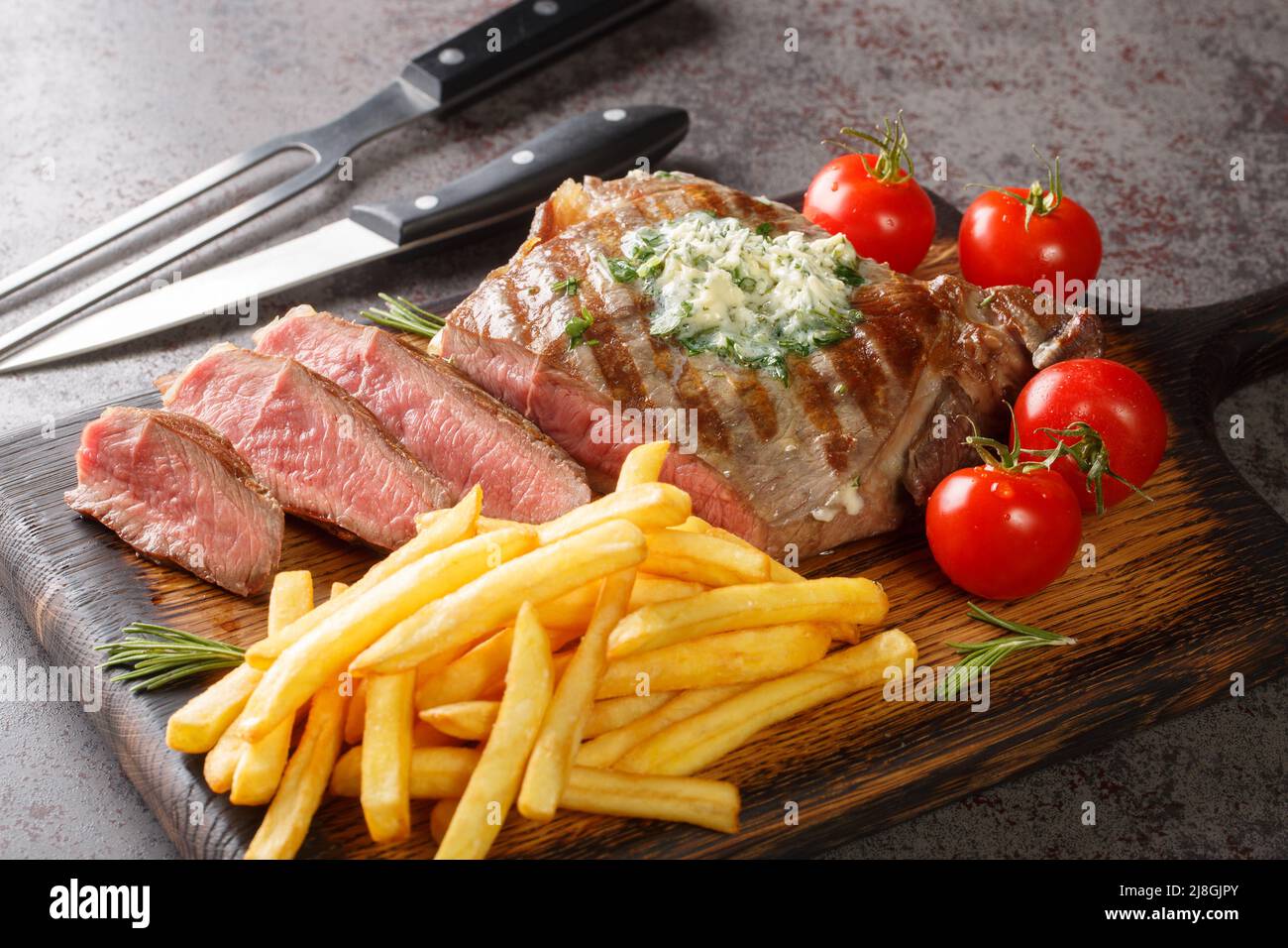 Freshly grilled steak with French Fries, green butter and tomatoes closeup on the wooden board on the table. Horizontal Stock Photo