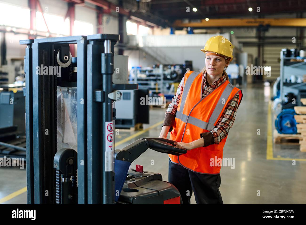 Young female engineer in protective helmet and uniform driving electric forklift while moving along warehouse or factory workshop Stock Photo