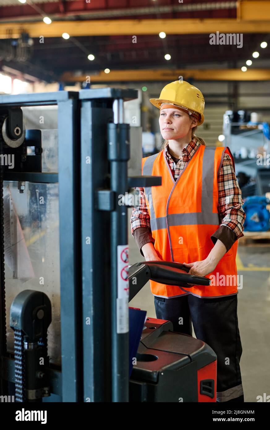 Female engineer looking forwards while standing in front of electric forklift and moving along aisle of workshop or warehouse Stock Photo