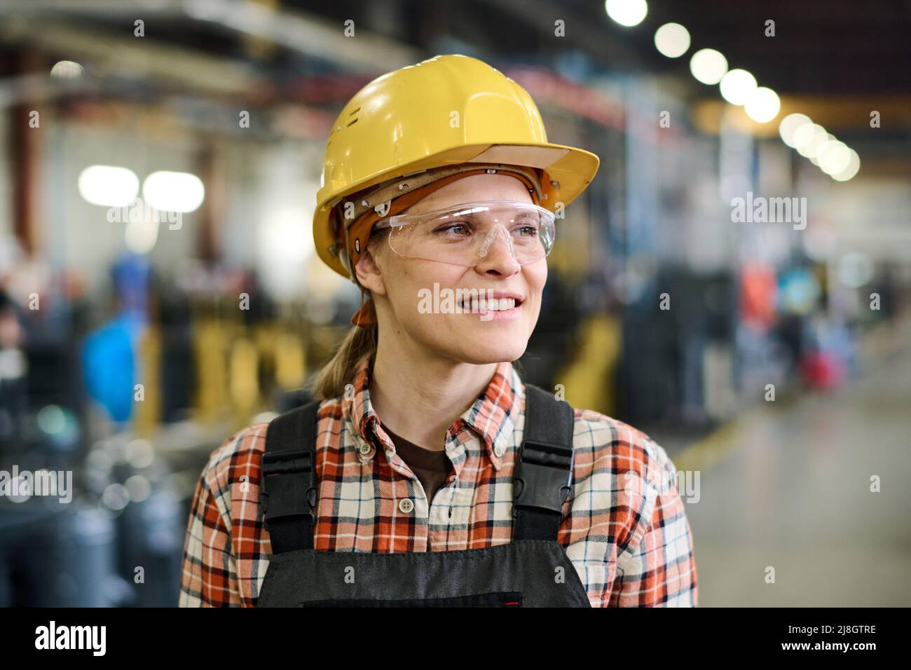 Happy young female engineer in hardhat and coveralls standing in workshop or warehouse of factory with industrial equipment Stock Photo