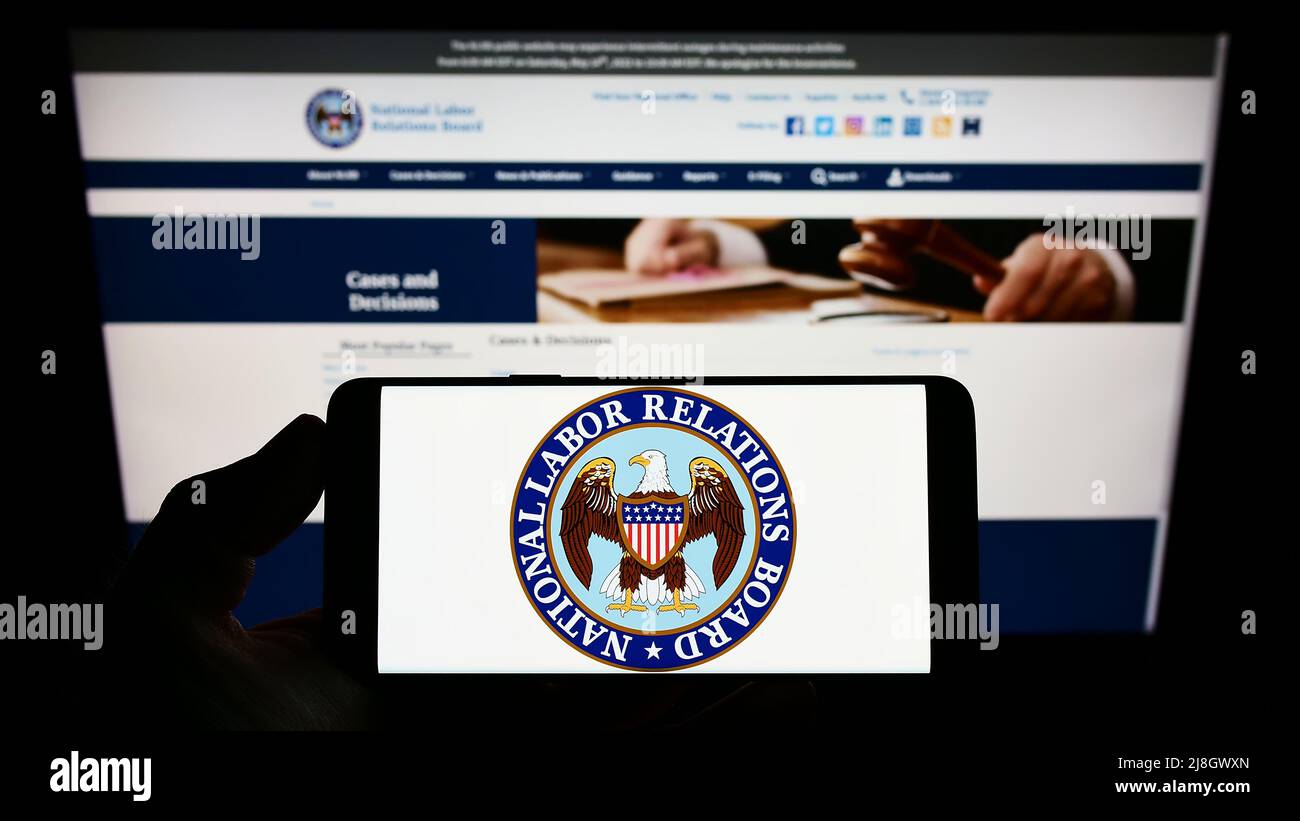 Person holding cellphone with logo of US National Labor Relations Board (NLRB) on screen in front of webpage. Focus on phone display. Stock Photo
