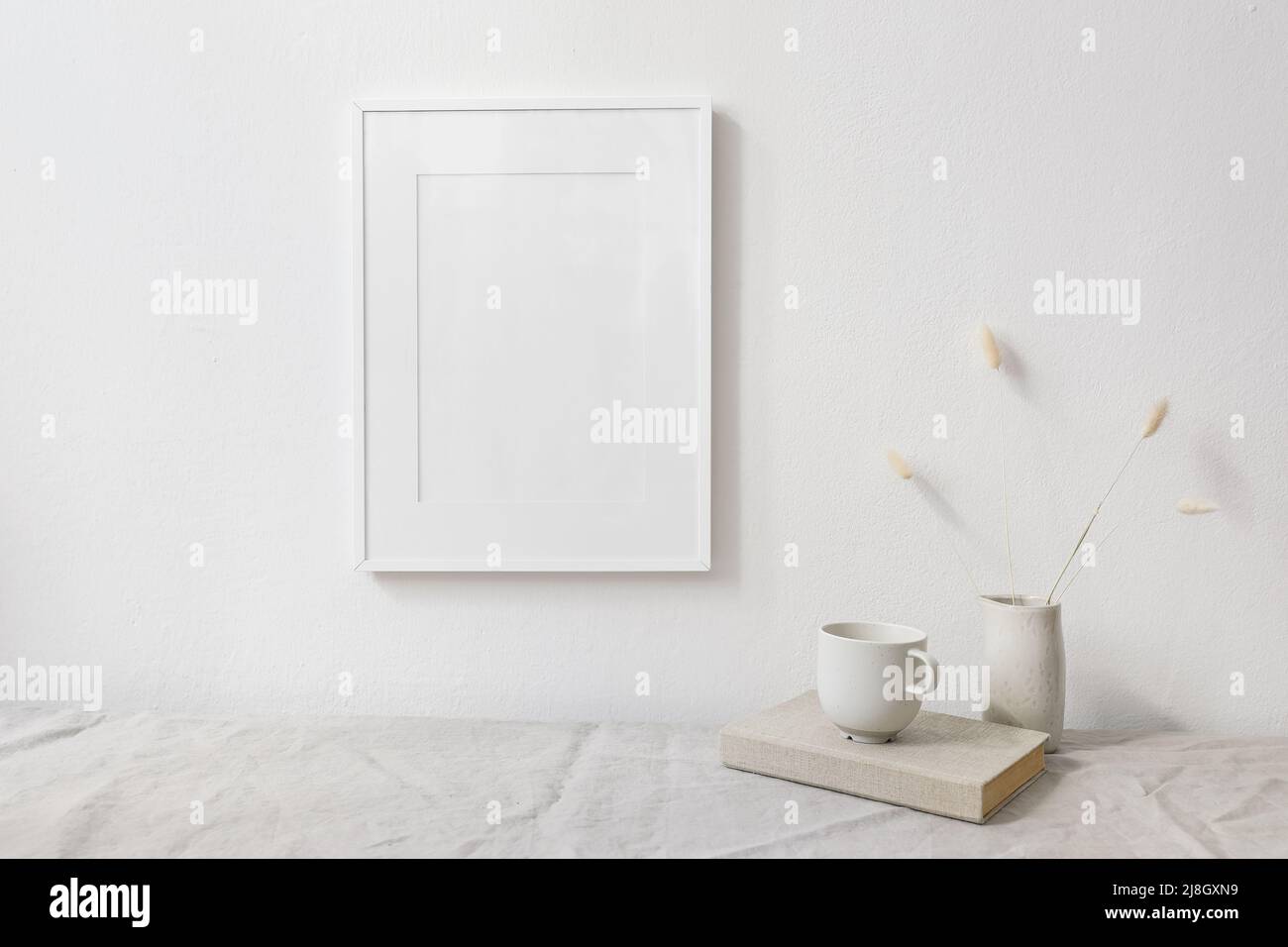 Neutral breakfast still life scene. White wooden picture frame mockup. Vase with dry lagurus bunny tail grass. Cup of coffee on book. Linen table Stock Photo