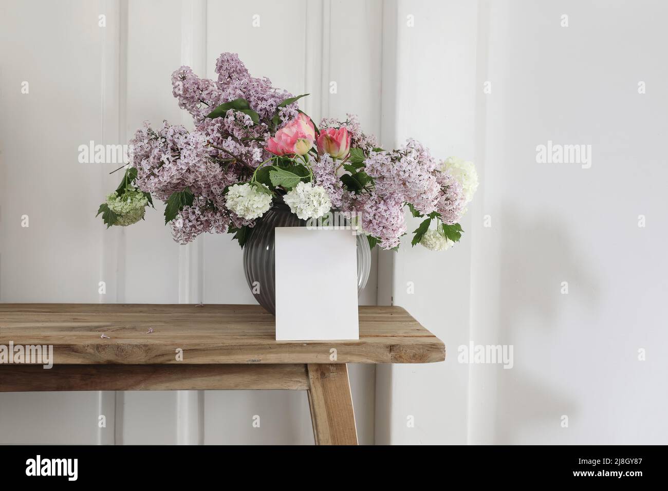 Vintage floral stationery mockup. Blank greeting card, invitation. Vase with bouquet of lilac, viburnum and tulips on old wooden stool. White wooden Stock Photo