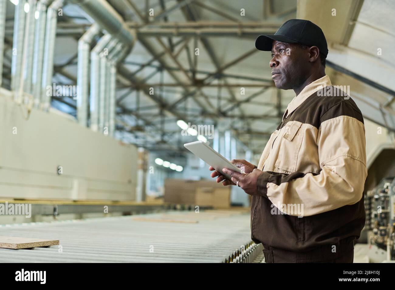 Serious African construction worker in uniform working on digital tablet while standing in workshop of factory Stock Photo